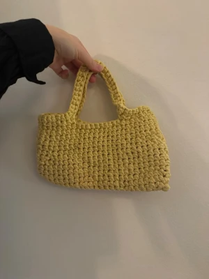 Gul virkad handväska - Säljer en söt gul handväska som är virkad med grova maskor. Väskan har två handtag och en mjuk, lite boxig form. Perfekt accessoar för dig som vill ha något unikt och färgglatt till din outfit.