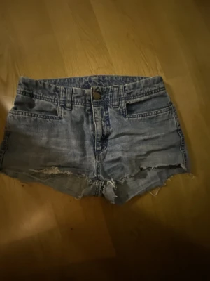 Låg midjade blå jeansshorts  - Säljer ett par klassiska blå jeansshorts med råa, fransiga kanter och slitningar framtill. Shortsen har fem fickor, bälteshällor och knappgylf. Perfekta för varma dagar och en avslappnad look.
