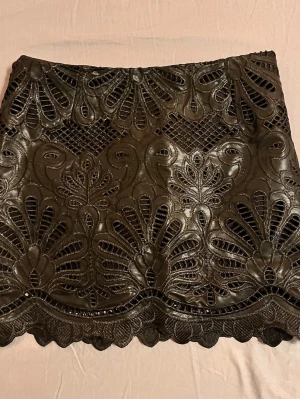 Svart kortkjol med spetsdetaljer - Supersnygg svart kortkjol med broderade spetsdetaljer och coolt mönster. Kjolen har en vågig kant nertill och ett genomarbetat, nästan blommigt mönster som ger en unik look. Perfekt för dig som vill sticka ut lite extra.