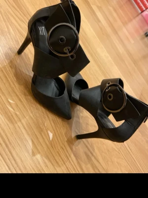 Svarta pumps med stor metallring XIT - Säljer ett par svarta pumps från XIT med spetsig tå och hög klack. Skorna har breda band över foten och en stor dekorativ metallring som detalj. Materialet är matt och känns som syntet. Dragkedja bak för enkel påtagning.