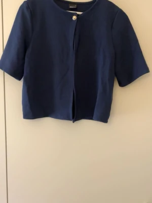 Mörkblå kofta med kort ärm - Säljer en stilren mörkblå kofta från Selected Femme. Koftan har korta ärmar, rund halsringning och stängs med en dekorativ knapp framtill. Perfekt att slänga över en topp för en clean look.