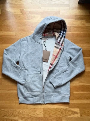  Burberry Grå Zip Hoodie med Check-foder• Burberry Hoodie Grå – Zip & Klassisk Check• Burberry Zip Hoodie Grey –  Original• Burberry Grå Hoodie med Dragkedja -  Grå zip-hoodie från Burberry i mjukt och bekvämt material. Klassiskt rutigt foder (Burberry check) på insidan av huvan och längs dragkedjan. Hel dragkedja framtill, två framfickor och ribbade muddar i ärmslut och nederkant. Stilren och tidlös modell som passar till vardags. Mycket fint skick, originaltag kvar