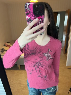 Rosa långärmad topp med stjärndetalj - Säljer en rosa långärmad topp med svart abstrakt tryck och en glittrig stjärna av små nitar på bröstet. Rund halsringning och mjukt material som känns skönt mot huden. Perfekt för dig som gillar lite extra detaljer på vardagslooken.