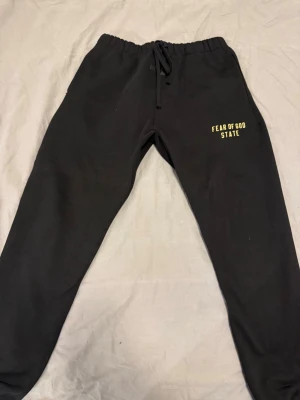 Svarta Fear of God Essentials mjukisbyxor - Svarta mjukisbyxor från Fear of God Essentials med gul text 'FEAR OF GOD STATE' på vänster ben. Byxorna har dragsko i midjan och är tillverkade i en mjuk bomullsblandning med polyester. Perfekta för chill dagar och streetwear-vibe.