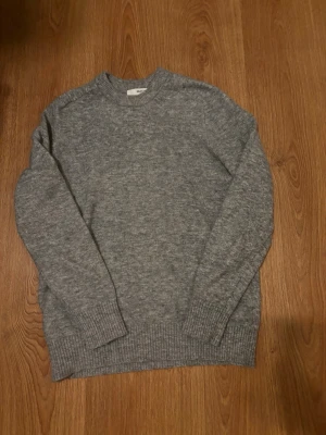 Grå stickad tröja från Selected Homme - Mysig grå stickad tröja från Selected Homme i klassisk modell. Tröjan har rund halsringning, ribbade muddar vid ärmslut och nederkant samt lång ärm. Perfekt för lager på lager under kyliga dagar.