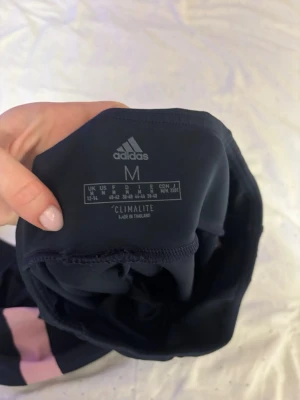 Svarta Adidas leggings med rosa detaljer - Säljer ett par svarta Adidas leggings i storlek M med snygga rosa detaljer vid benen. Tillverkade i funktionsmaterialet Climalite som andas och passar perfekt till träning. Tight passform och hög midja för skön känsla och stilren look.