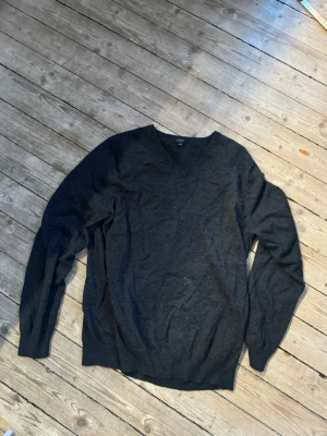 Mörkgrå stickad tröja från J.Crew - Snygg mörkgrå stickad tröja från J.Crew i storlek L. Tröjan har v-ringning och långa ärmar, perfekt för lager på lager under kyliga dagar. Enkel och stilren design som funkar till det mesta.