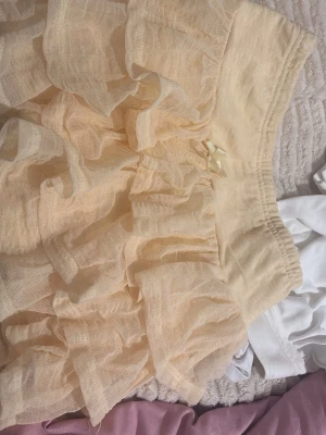 Beige volangkjol med rosett och shorts under  - Supersöt beige kortkjol med flera lager volanger och en liten rosett framtill i midjan. Kjolen har elastisk midja och är gjord i ett tunt, lätt material som ger en fluffig look. Perfekt för dig som gillar romantisk och lekfull stil.