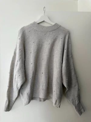 Grå stickad tröja med pärlor H&M - Mysig grå stickad tröja från H&M med silverfärgade pärlor över hela tröjan. Tröjan har en ribbad, hög halsringning och långa ärmar med breda muddar. Perfekt för dig som gillar en enkel men ändå lite extra look.