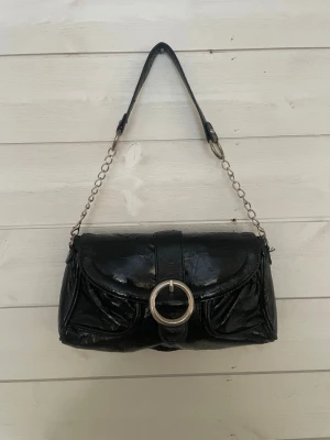 Svart axelväska med kedjedetaljer, vintagde  - Snygg svart axelväska i glansigt skinnimitation med silverfärgad kedja och stor rund metallring framtill. Väskan har ett klassiskt format lock och kort axelrem i kombination av kedja och skinn. Perfekt för dig som gillar en stilren men edgy look. Köpt för flera år sedan på vintagde secondhand 