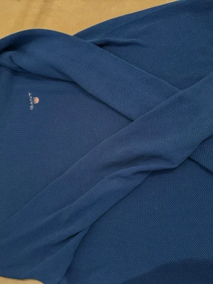 Blå stickad tröja från GANT - Säljer en blå stickad tröja från GANT i bomull. Tröjan har rund halsringning, diskret GANT-logga med sköld på bröstet och ribbade muddar. Perfekt till lager på lager och enkel att matcha med jeans eller chinos.