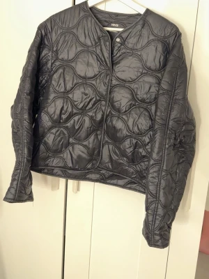 Svart quiltad jacka från MNG - Säljer en svart quiltad jacka från MNG i storlek M. Jackan har rund halsringning, tryckknappar framtill och ett vågigt quiltat mönster. Materialet är glansigt och lätt, perfekt för lager på lager under höst och vår.