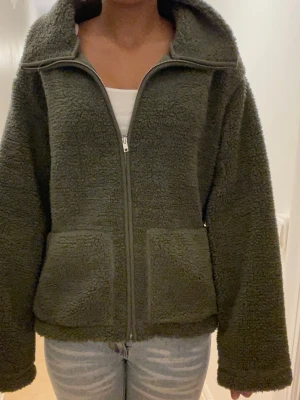 Olivgrön teddyjacka med dragkedja - Mysig olivgrön teddyjacka med stor krage och två stora fickor framtill. Jackan har hel dragkedja och är tillverkad i fluffigt fleece-material!💚