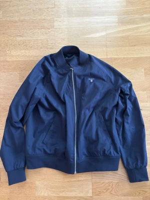 Marinblå bomberjacka från GANT - Snygg marinblå bomberjacka från GANT med klassisk ribbad krage och muddar. Jackan har dragkedja framtill, två snedställda fickor och diskret GANT-logga broderad på bröstet. Perfekt för dig som gillar stilren och enkel design.