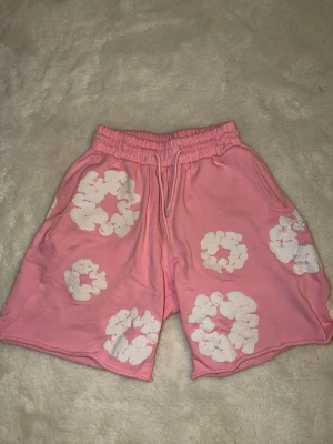 Denim tears shorts - Säljer ett par rosa shorts med elastisk midja och snörning. Shortsen har ett unikt blommigt mönster i vitt över hela plagget och är tillverkade i mjuk bomull. Perfekta för sommarens chill dagar och har en loose fit för extra komfort.