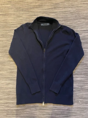 Marinblå kofta från Milano - Klassisk marinblå kofta från Milano med hel dragkedja framtill och ribbad krage. Tillverkad i mjuk bomullsmix med lite elastan för extra komfort. Perfekt till lager på lager och har långa ärmar samt ribbade muddar. Om ni har några frågor så är det bara att höra av er!😊