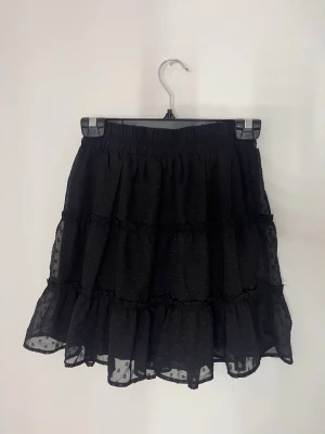 Svart volangkjol med prickigt mönster - Supersöt svart kortkjol med volanger och diskret prickigt mönster. Kjolen har resår i midjan och är gjord i ett lätt, transparent tyg med flera lager för extra volym. Kan bäras som hög & låg midjat. 