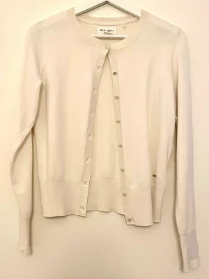 Naturvit kofta från Holly & White - Säljer en stilren naturvit kofta från Holly & White. Koftan har rund halsringning, långa ärmar och knäppning framtill med små knappar. Perfekt att slänga över ett linne eller t-shirt för en enkel och snygg look.