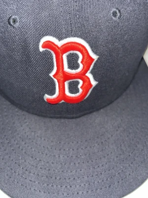 Boston Red Sox New Era keps - Säljer en mörkblå New Era 59FIFTY keps med Boston Red Sox logga broderad i rött och vitt framtill. Klassisk MLB-logga baktill och rak skärm. Perfekt för dig som vill ha en clean och sportig look.