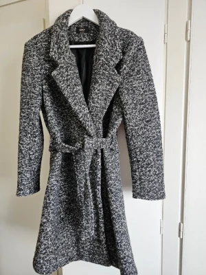 Gråmelerad kappa från ONLY med bälte - Säljer en gråmelerad kappa från ONLY i storlek L. Kappan har ett snyggt bälte i midjan, bred krage och är helfodrad. Materialet är en mix av polyester sopch ull, vilket gör den både varm och stilren. 