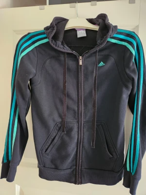 Adidas svart hoodie. storlek S - Svart Adidas hoodie med dragkedja och turkosa ränder på ärmarna. Den har en luva med dragsko och två fickor fram.
