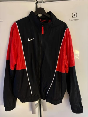 Nike vindjacka svart/röd L - Säljer en riktigt snygg vindjacka från Nike i svart och rött med vita detaljer. Jackan har dragkedja framtill, meshfoder och klassisk Nike-logga på bröstet. Perfekt för dig som vill ha sportig stil och bekväm passform.