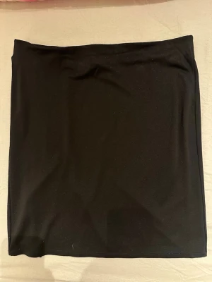 Svart kortkjol från Monki - Enkel svart kortkjol från Monki i storlek L. Kjolen har en rak passform och är gjord i mjukt stretchigt material med en liten slits baktill. Perfekt basplagg till garderoben.Har används en gång och har mycket bra skick 