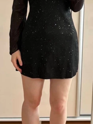 Svart glittrig kortklänning - Säljer en svart kortklänning med glittrigt mönster. Klänningen har långärmade, lätt transparenta ärmar och en figurnära passform. Perfekt för dig som vill sticka ut med lite extra glitter. Supersnygg och trendig look!