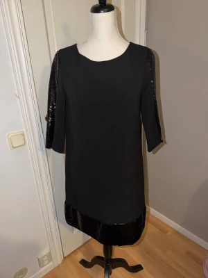 Svart paljettklänning från Stockh LM - Snygg svart kortklänning från Stockh LM med paljettdetaljer på ärmar och nederkant. Klänningen har rund halsringning, trekvartsärm och rak passform. Perfekt för dig som vill ha en stilren look med lite extra bling.