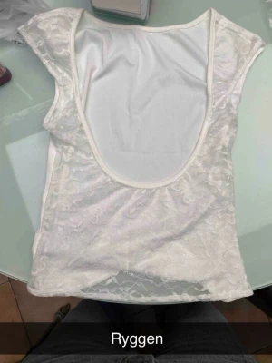 Vit topp med spets -  en fin vit topp med ribbad framsida och transparent spets med blommönster på ryggen och axlarna. Toppen har en djup, öppen rygg och är ärmlös med breda axelpartier. Sitter tight och är mycket stretchig den är M men passar S oxå skulle jag säga.