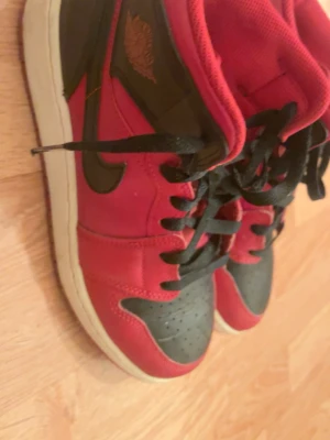 Nike Air Jordan 1 High Röd/Svart - Säljer ett par Nike Air Jordan 1 High i rött och svart med vita sulor. Skorna har klassisk snörning, svart swoosh och ikoniska Jordan-loggan på sidan. Ovandelen är i syntet och textil, med perforerad tåbox för extra ventilation. Perfekta för dig som gillar streetwear.