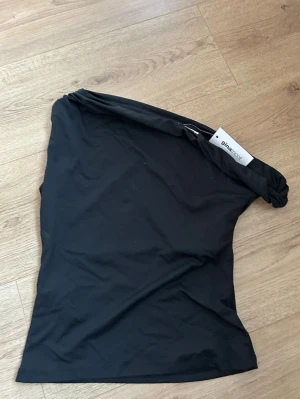 Svart oneshoulder topp från Gina Tricot - Säljer en svart oneshoulder topp från Gina Tricot. Toppen har en stilren och enkel design med en axel och är tillverkad i mjukt stretchigt material. Perfekt att styla med jeans eller kjol för en trendig look. Aldrig använd 