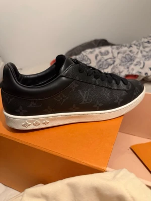 Louis Vuitton svarta sneakers i skinn - Louis Vuitton sneakers i svart skinn med klassiskt monogrammönster över hela skon. Skon har svarta skosnören, vit platt sula och rund tå. Märket syns på plösen och sulan. Snygg och tidlös modell som passar till det mesta.