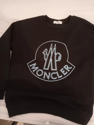 Svart sweatshirt från Moncler - Säljer en svart sweatshirt från Moncler med stort vitt logotryck på bröstet. Tröjan har rund halsringning och långa ärmar. Perfekt för dig som vill ha en clean och stilren look med exklusiv känsla.