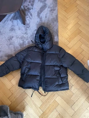 Svart pufferjacka från H&M XL - Säljer en svart pufferjacka från H&M i storlek XL. Jackan har huva med snörning, dragkedja framtill och stora vadderade partier som ger en riktigt fet look. Perfekt för kalla dagar när du vill vara både varm och stilren.