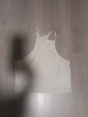 Vit ribbad linnetopp - Säljer en enkel och stilren vit linnetopp med smala axelband och hög halsringning. Toppen är ribbad och croppad, perfekt till sommaren eller för att styla lager på lager. Passar till många olika looks.