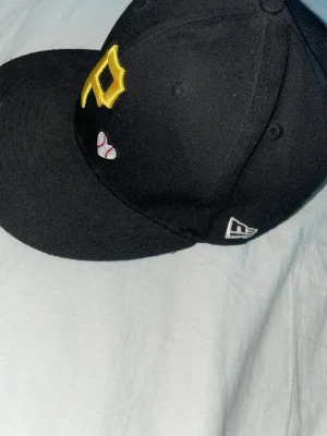 Svart New Era keps med gul detalj - Säljer en svart keps från New Era med gul broderad bokstav på framsidan och en liten baseboll-detalj. Klassisk rak skärm och vit New Era-logga på sidan. Perfekt för dig som gillar sportig stil och vill ha en clean accessoar.