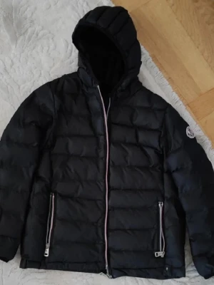 Svart Moncler dunjacka med huva - Säljer en svart Moncler dunjacka med huva och broderad logga på ärmen. Jackan har två fickor med dragkedja och en snygg quiltad design. Perfekt för kalla dagar och riktigt stilren look. Har några enstaka hål inget som man ser när man har på sig den. Dragkedja är sönder men går att lagas 