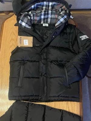 Svart pufferjacka från Burberry - Säljer en svart pufferjacka från Burberry med klassiskt rutigt foder i huvan. Jackan har dragkedja, knappar och två fickor med dragkedja. Oversized look och riktigt skön för kalla dagar. Märket är broderat på ärmen och materialet är polyester.