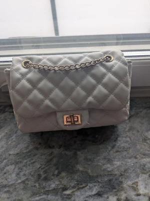 Vit quiltad axelväska med kedja - Säljer en stilren vit axelväska med quiltat mönster och guldfärgad kedja. Väskan har ett vridlås i metall och mjukt syntetmaterial. Perfekt storlek för det viktigaste och ger en classy vibe till din outfit. Nypris 199