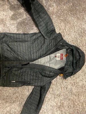 Mörkgrön lättviktsjacka från Parajumpers - Säljer en mörkgrön quiltad lättviktsjacka från Parajumpers med huva och flera dragkedjeförsedda fickor. Jackan har en snygg patch på ärmen och orange detalj vid halsen. Perfekt för dig som vill ha en smidig och stilren jacka till vardagen.
