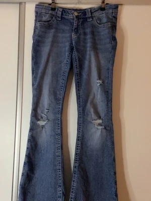 Blå bootcut jeans med broderad ficka - Säljer ett par blå bootcut jeans med slitningar på benen och snyggt broderad bakficka med vinge. Jeansen har klassisk femficksmodell, normal passform och är tillverkade i mjukt denimtyg. Perfekta för dig som gillar detaljer och en avslappnad look.