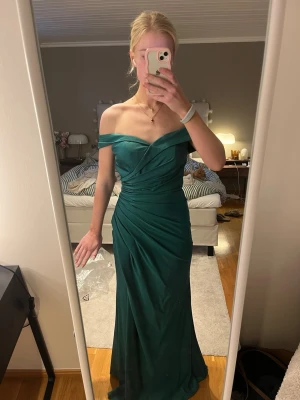Grön axelbandslös långklänning - Säljer en elegant smaragdgrön långklänning med offshoulder och draperad överdel. Klänningen har en snygg slits framtill och figurnära passform som framhäver siluetten. Perfekt för bal eller festliga tillfällen. Använd en gång. Köpt på Remanns i Göteborg för ca 2500kr. Har en minimal fläck på insidan av byst (se bild) därav lägre pris, annars i super bra skick. Kupan är en B/C kupa. Längd från framsida byst till slutet av kjolen är 137