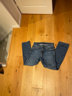 Mörkblå jeans från Nudie Jeans - Säljer ett par klassiska mörkblå jeans från Nudie Jeans med raka ben och femficksmodell. Jeansen har snygga kontrastsömmar och Nudie-loggan på bakfickan. Tillverkade i slitstark bomull med en tidlös look som passar till allt.