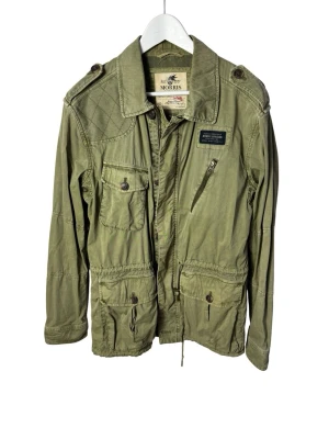 Olivgrön parkajacka från Morris - Säljer en olivgrön parkajacka från Morris med flera fickor, axelklaffar och snygga detaljer som dragkedja och knappar. Jackan har justerbar midja med snörning och quiltad axel. Perfekt för dig som gillar utility-vibe och vill ha en stilren men funktionell jacka.