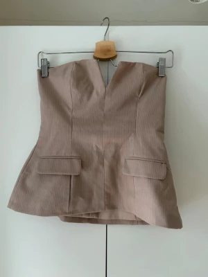 Beige randig korsettopp - Säljer en beige korsettopp med tunna vita ränder och figurnära passform. Toppen är axelbandslös med en snygg v-ringning framtill och dekorativa ficklock. Perfekt att styla med jeans eller kjol för en trendig look.