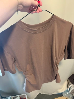 Figursydd t-shirt  - Figursydd brun t-shirt i mjuk bomull med rund hals och korta ärmar. Modellen är rak och har en liten slits bak för extra rörelsefrihet. Perfekt för en chill och avslappnad stil.