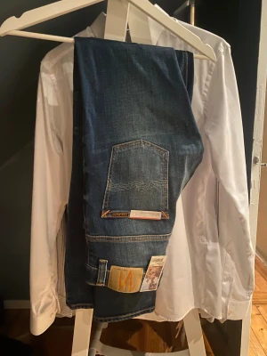 Nudie Jeans blå denimjeans - Säljer ett par klassiska blå jeans från Nudie Jeans med orangea sömmar och snygga detaljer på bakfickan. Jeansen har en rak passform och är tillverkade i denim. Perfekta för dig som gillar stilrena och tidlösa jeans med schysst tvätt. De är dessutom HELT NYA! Storlek: W30 L34