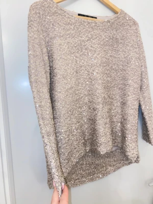 Beige glittrig stickad tröja - Säljer en beige stickad tröja med glittriga paljettdetaljer. Tröjan har långärmad design och en rund halsringning. Materialet är mjukt och har en snygg struktur som ger extra shimmer. Perfekt för dig som gillar att sticka ut med lite glitter i vardagen.