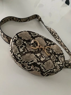 Axelväska med ormmönster från Tally Weijl - Snygg oval axelväska från Tally Weijl med ormmönster i beige och svart. Väskan har guldfärgade detaljer framtill och justerbar axelrem. Stängs med dragkedja och har ett rymligt fack. Perfekt för dig som vill sticka ut med coola detaljer.